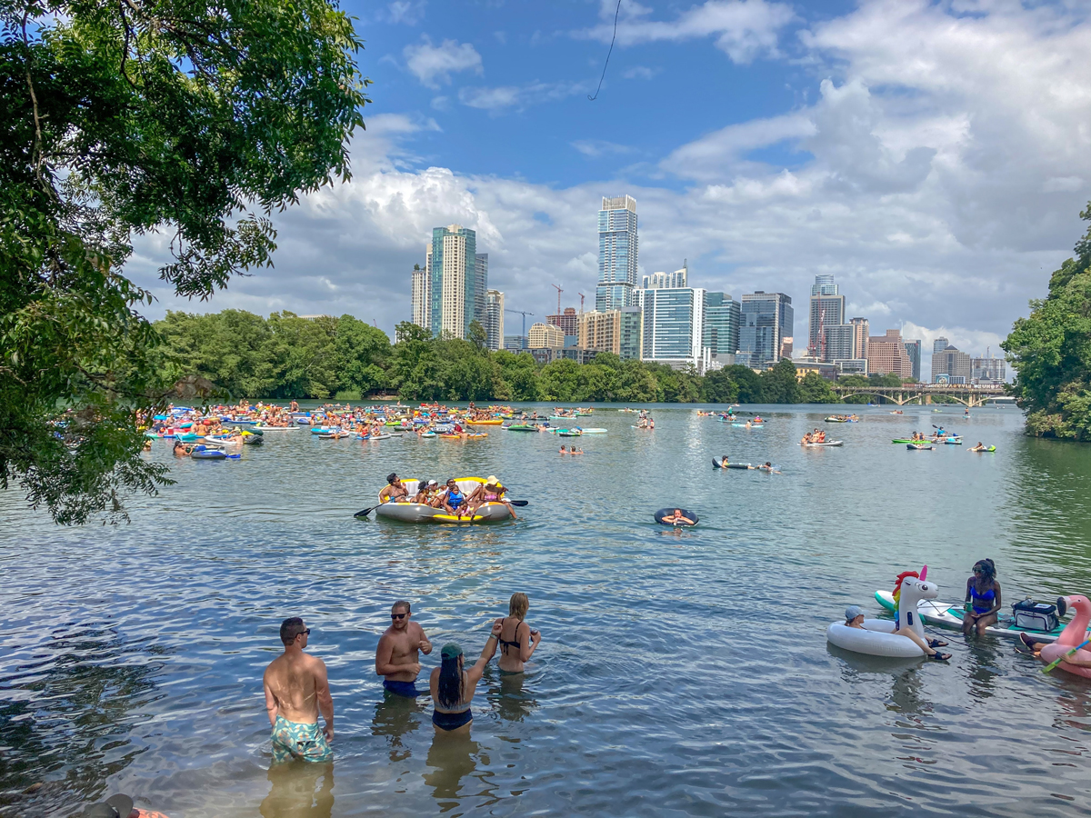 Lots of people floating on inflatable boats on a hot day in the town lake in Austin with the city skyline in the background. 