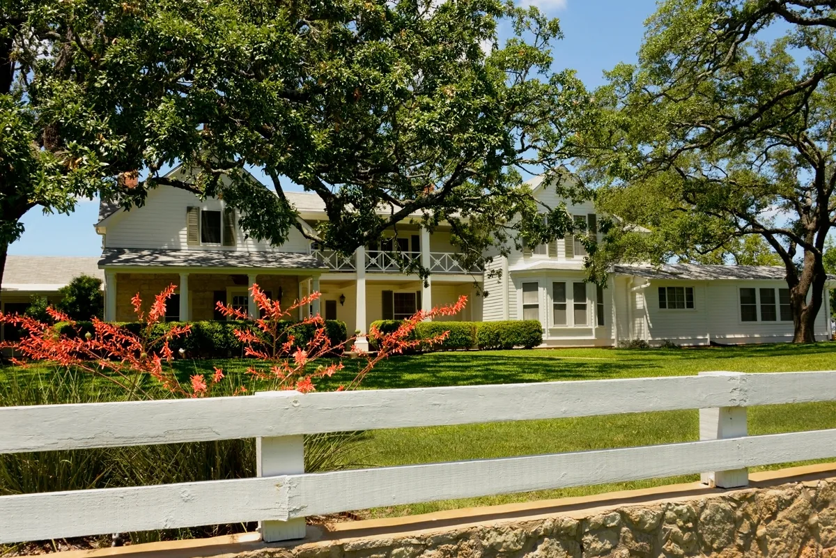 The white ranch house of President LBJ Johnson with green lawn and flowers along a white wooden fence. 