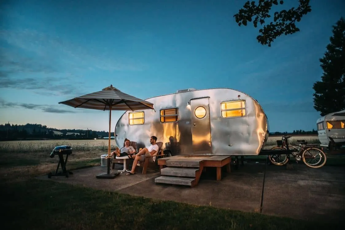 Couple sitting in front of a silver retro caravan at a campsite.