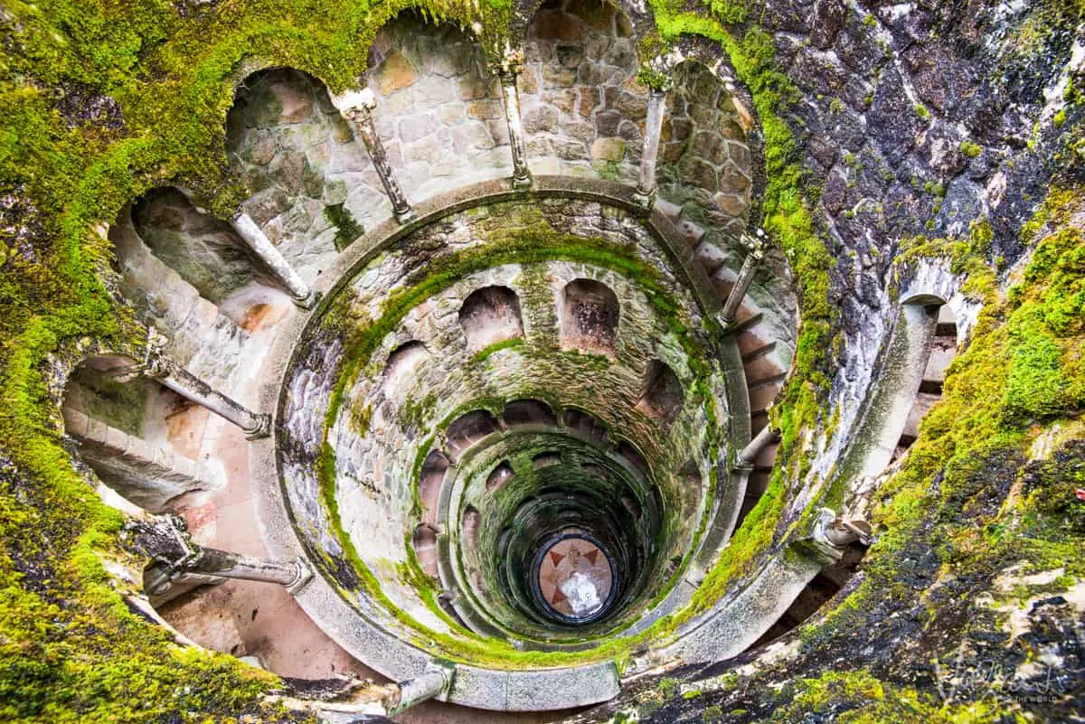 A deep well covered in green moss with an internal intricate winding staircase. It is the Initiation Well at Quinta da Regaleira in Sintra Portugal. 