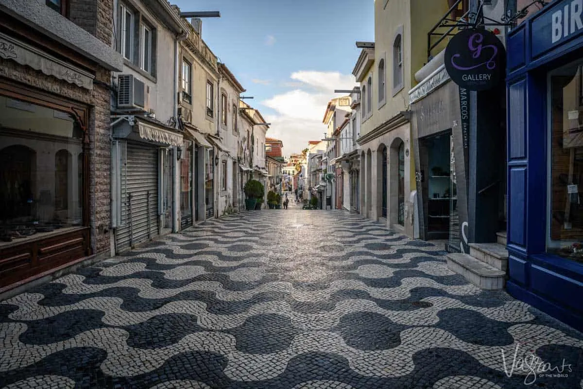 A typical shopping street in the town of Cascais in Portugal where the street is lined with the black and white patterned cobble stones that a typical in Portugal. 