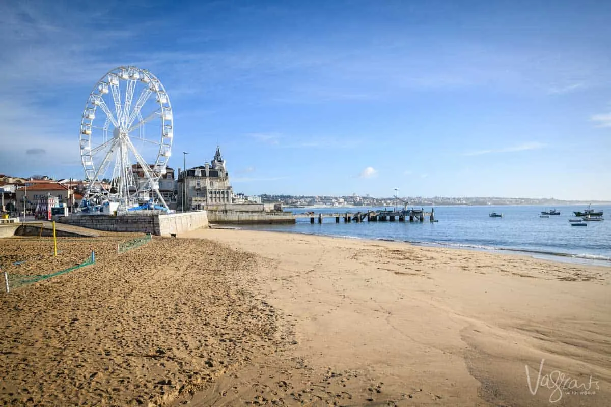 The beach front of Cascais in Portugal with a ferris wheel on the beach and a historical monastery building in the distance..