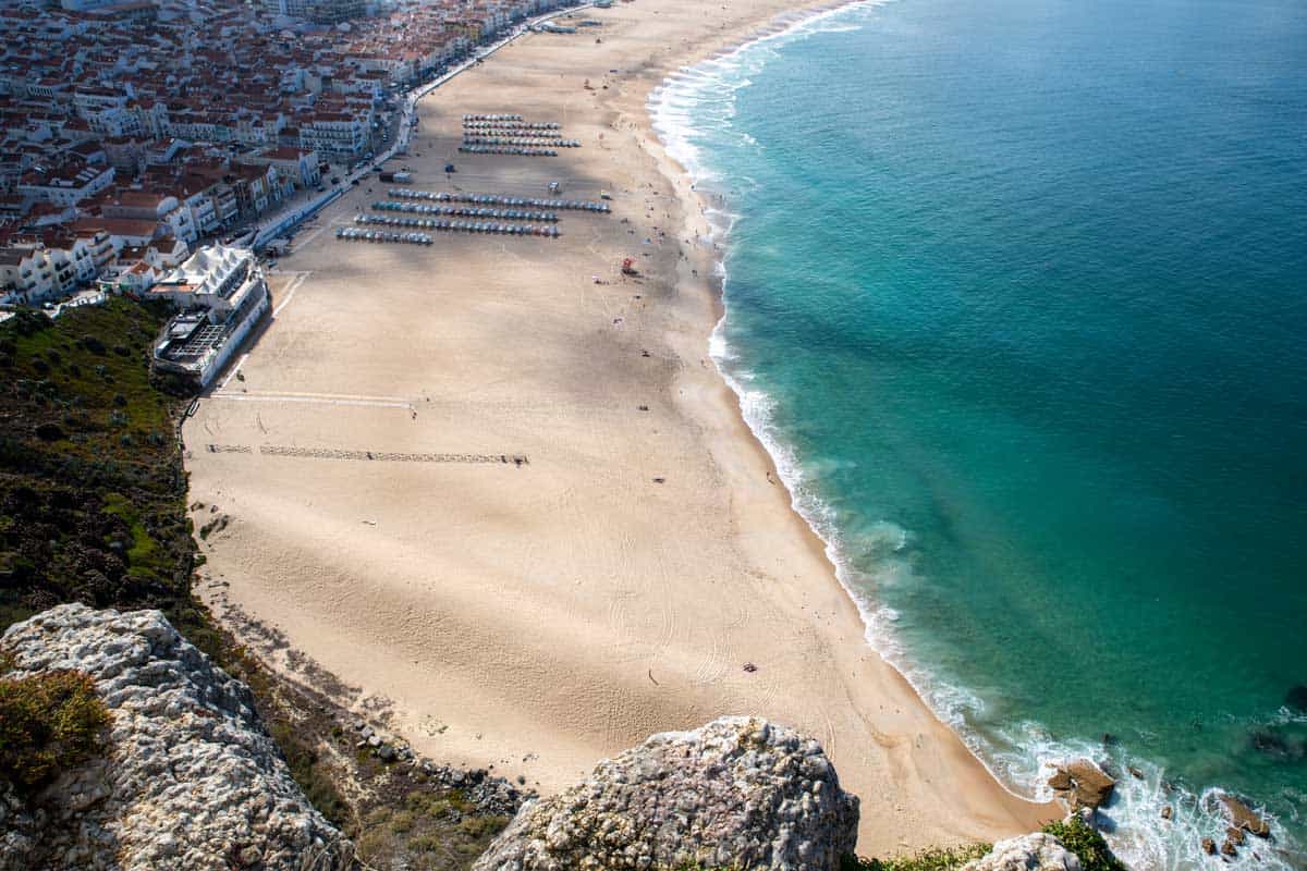 Looking over Praia de Nazaré in Portugal. The beach town spreads from the wide white beach with white building with red roofs. There is a few rows of organised beach chairs on the beach. 