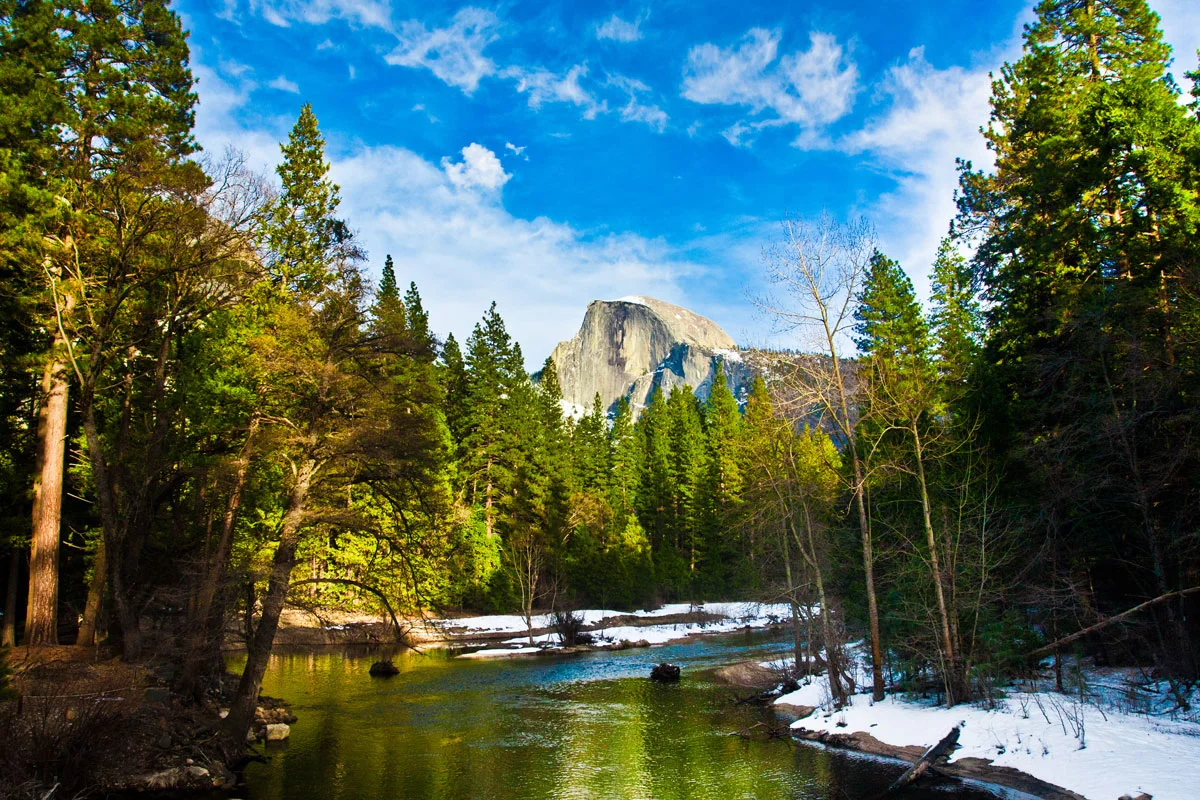 View of Half Dome down the river in forest with snow on the banks in Yosemite NP. 