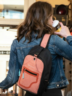 Back of a woman in a cafe drinking takeaway coffee wearing a pink sling backpack.