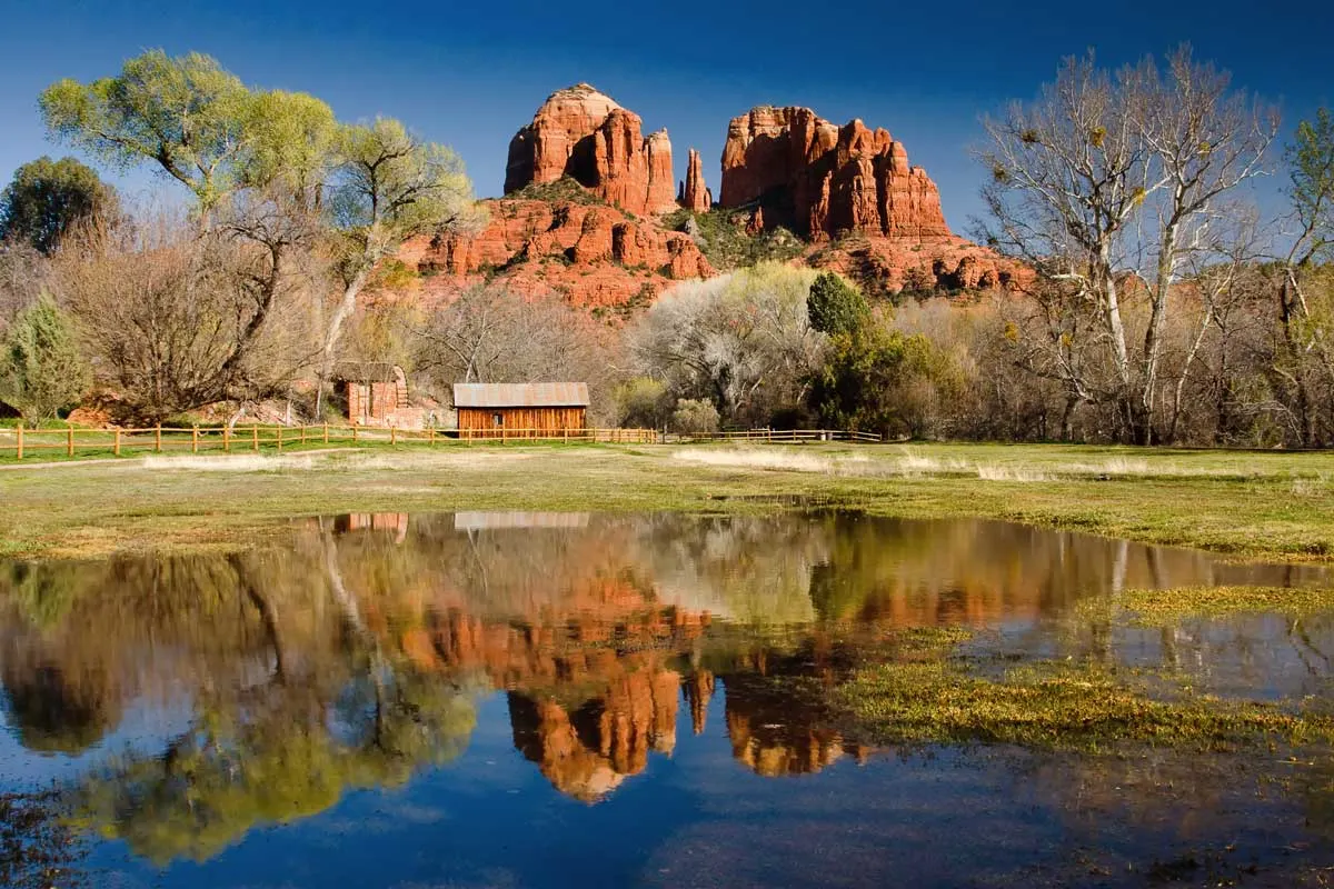 Red rocks of Cathedral Rock Sedona reflected in a blue lake with a farm house on the banks. 