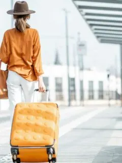 Female traveller pulling an orange suitcase weraing an orange shirt with white jeans and a large brown hat.