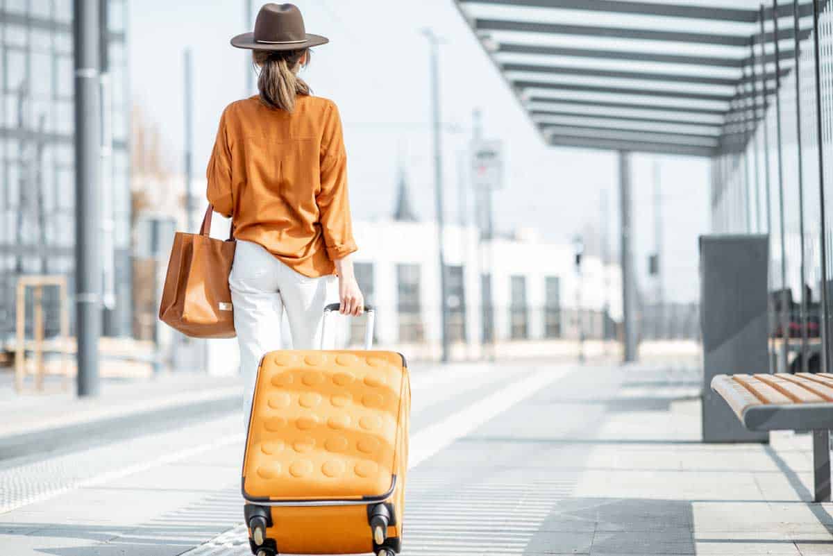Female traveller pulling an orange suitcase weraing an orange shirt with white jeans and a large brown hat.