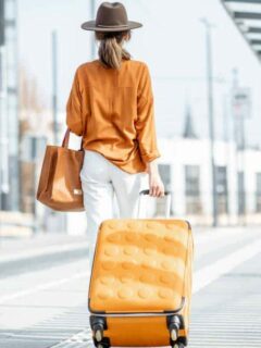Female traveller pulling an orange suitcase weraing an orange shirt with white jeans and a large brown hat.