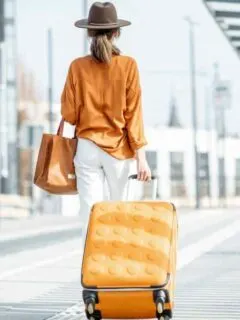Female traveller pulling an orange suitcase weraing an orange shirt with white jeans and a large brown hat.
