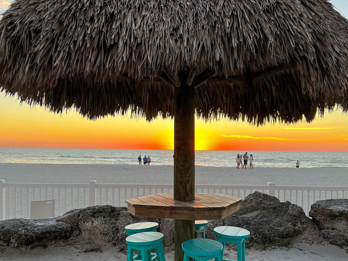 people watching a vibrant sunset from a white sand beach on Anna Maria Island in Florida. There is atypical tropical grass umbrella and bar stools in the foreground. 