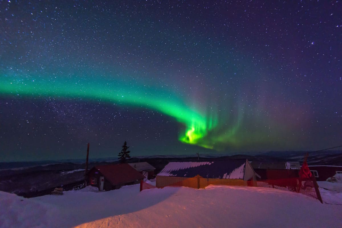 The gree northern lights light up the night sky over a house in a snowy landscape.