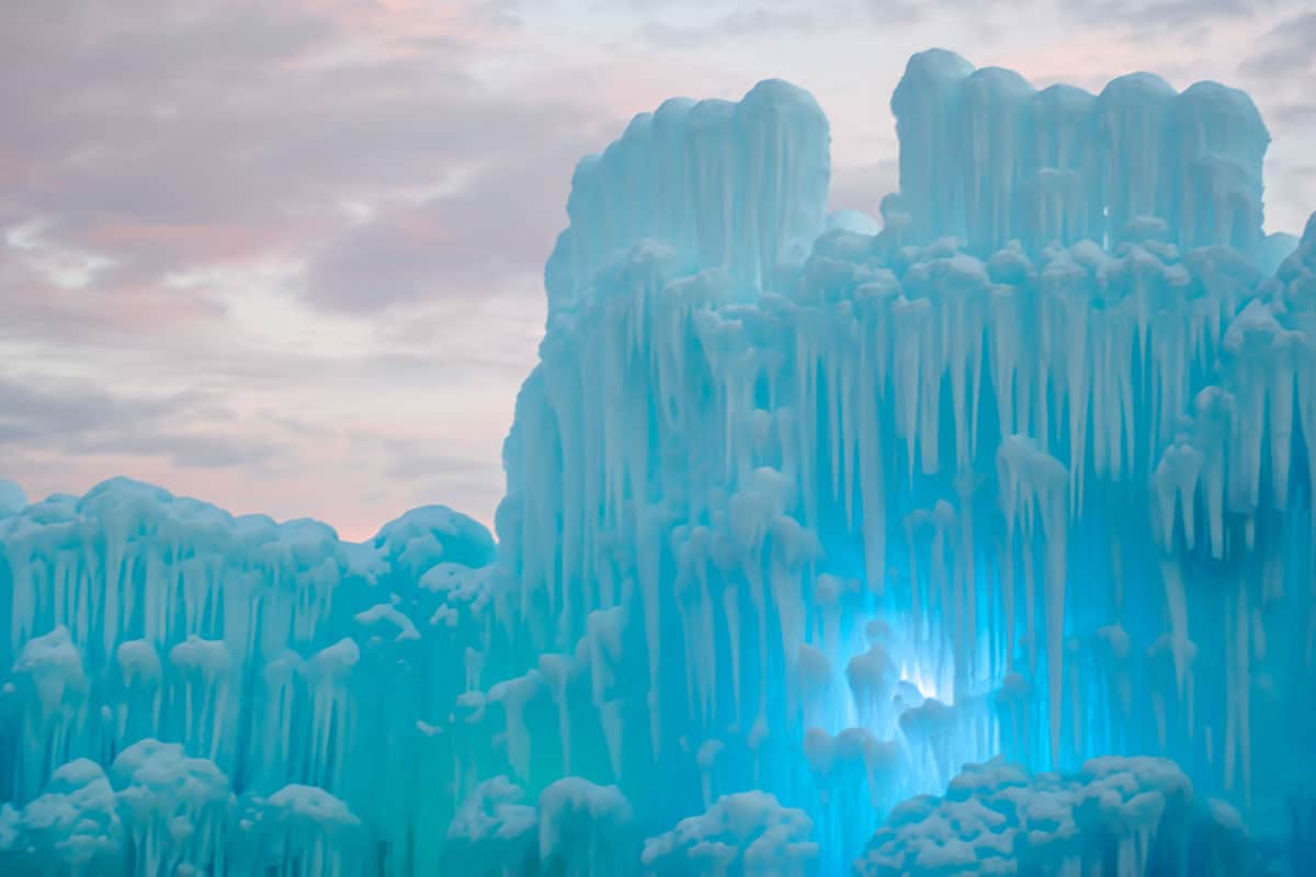 Man made ice castle glowing a light shade of blue with a pale pink sky in the background.