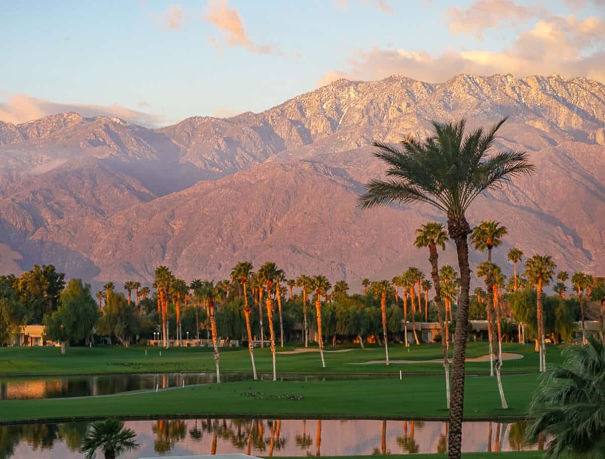 Sunset scene in Palm Springs with red mountains and lush green grass, lakes and palm trees.