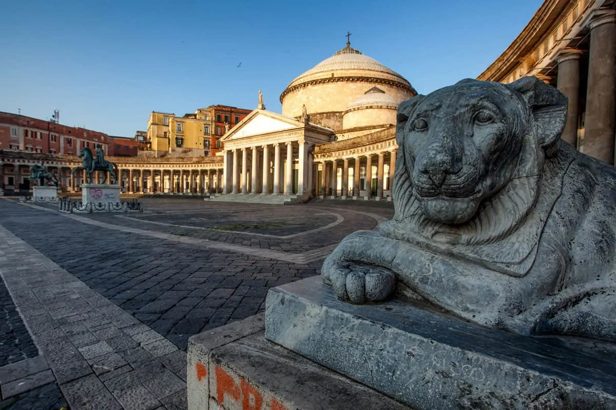 A lion statue in the shadows in Piazza Plebiscito in Naples. The domed buildings in the background are in golden sunset light. 