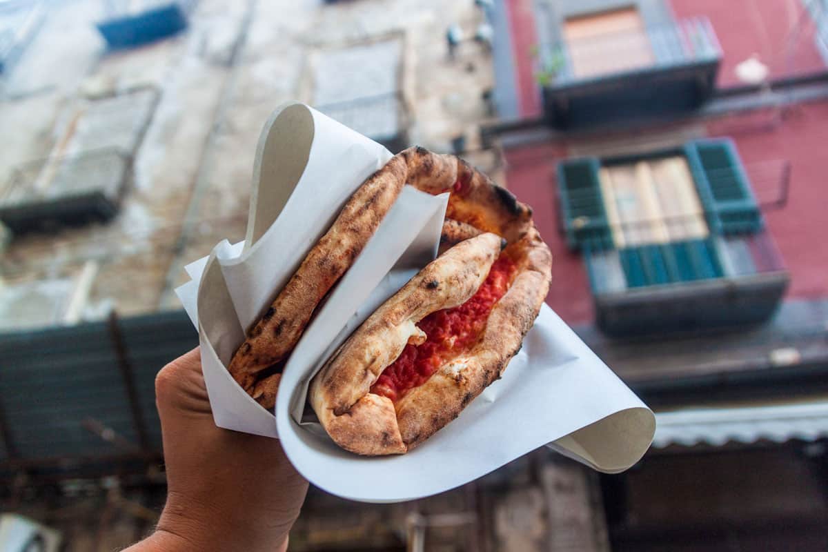 View of a hand holding a typical napoli pizza in the air in front of shuttered windows. The pizza is wrapped in white paper and folded.  