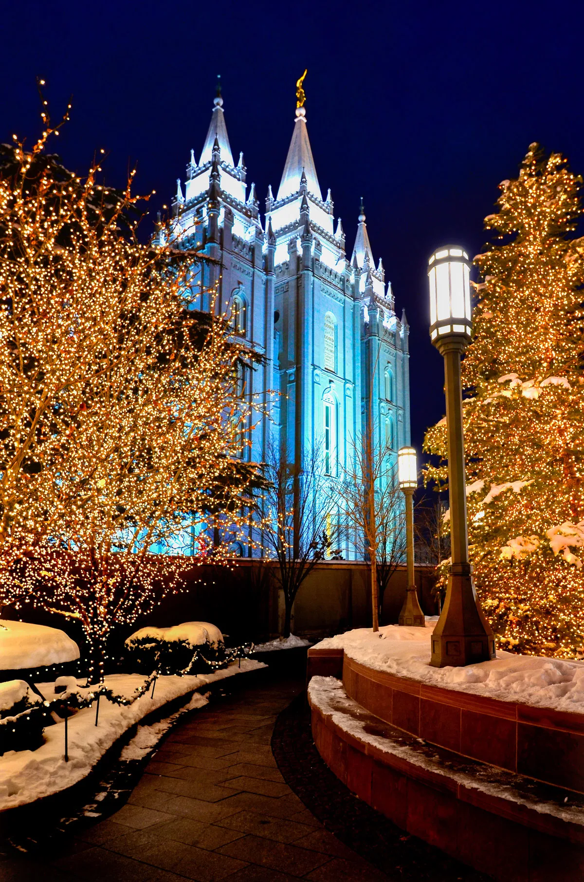 Salt Lake City Mormon Temple  on a snowy night lit up with Christmas lights and Christmas trees line the street. 
