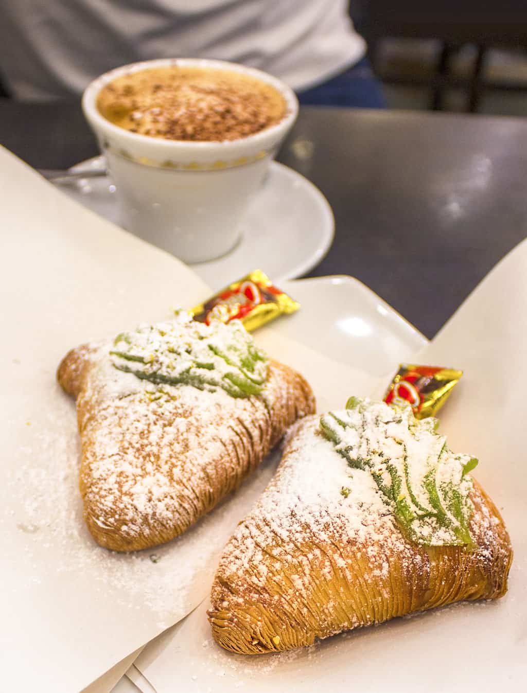 Two layered flaky pastries dusted with icing sugar called Sfogliatelle. They are a traditional pastry in Naples. There is also a cappuccino on the table.  