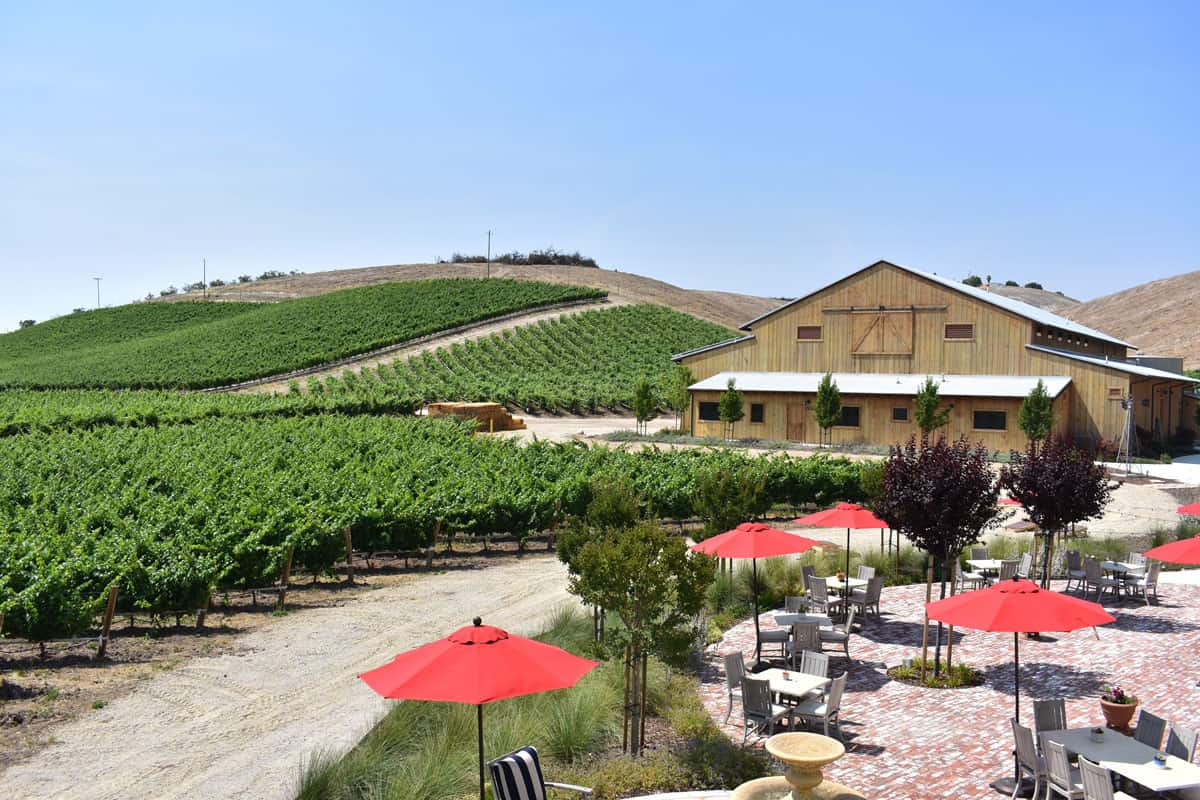 Red umbrellas open over outdoor restaurant tables at a winery in Paso Robles surrounded by green vineyards. 