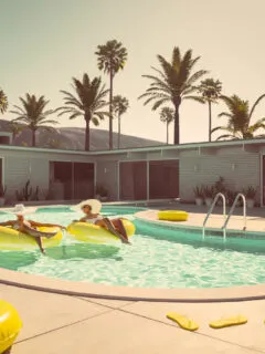 Two women floating a pool in Palm Springs on yellow tubes wearing big sun hats with palm trees in the background.