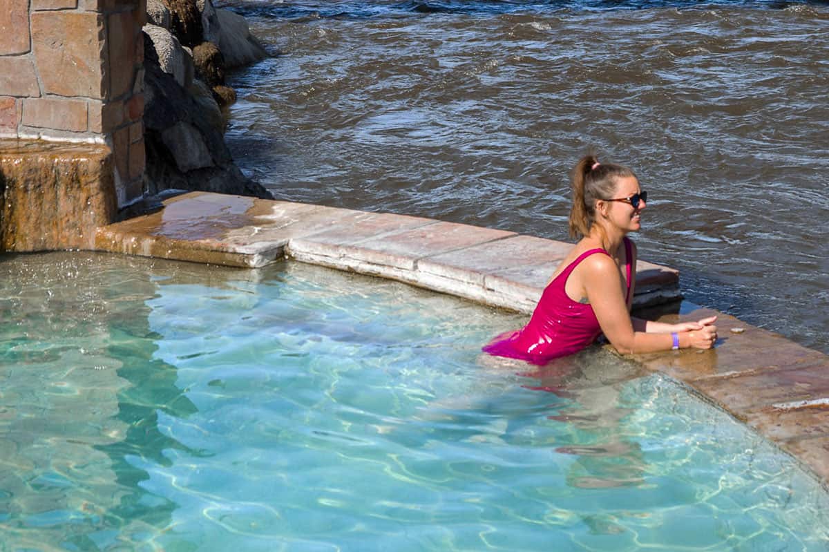 Lady in a pink bathing suit soaking in a hot spring pool overlooking a river. 
