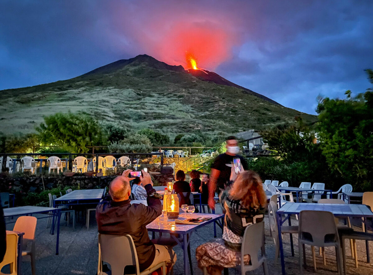 People sitting in an outdoor restaurant photographing a volcano spurting flames and lava in the distance on the Aeolian Islands in Italy. 