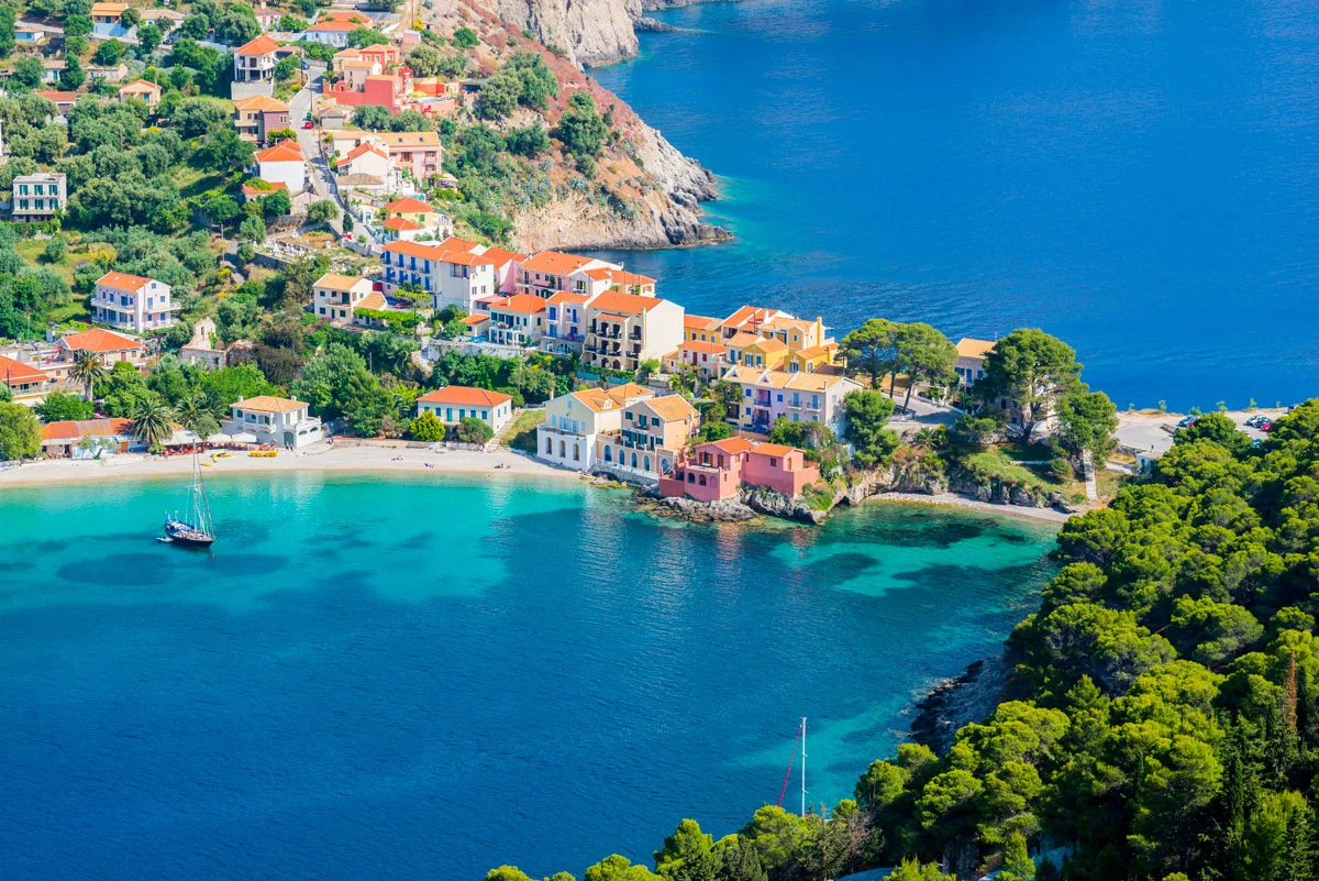 Colourful houses snake down the green hill and across a small strip of land diving to crystal blue bays of water. A white sand beach lines the side of the bay. 