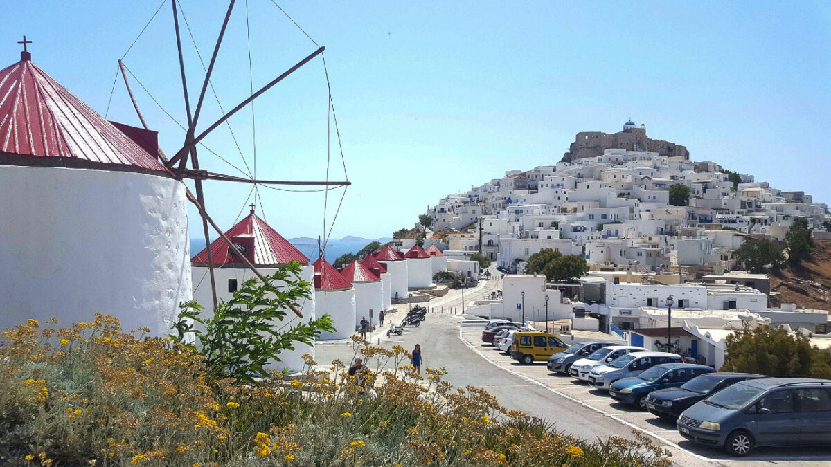 Traditional windmills in a row with a typical Greek Island village cascading down the hill in the distance. 