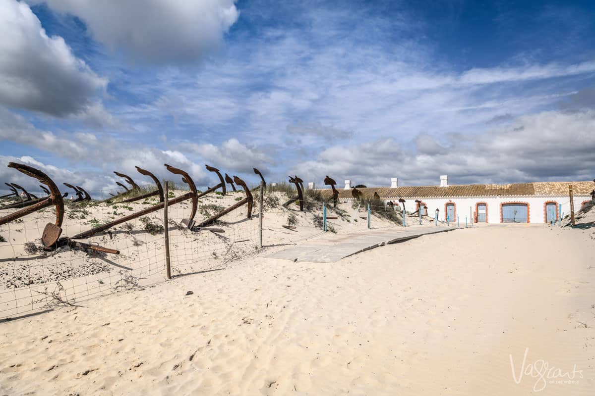Rusty anchors arranged in the white sand dunes with an old traditional stone fishermans building at the end of a wooden path on Barril Beach in Portugal. 