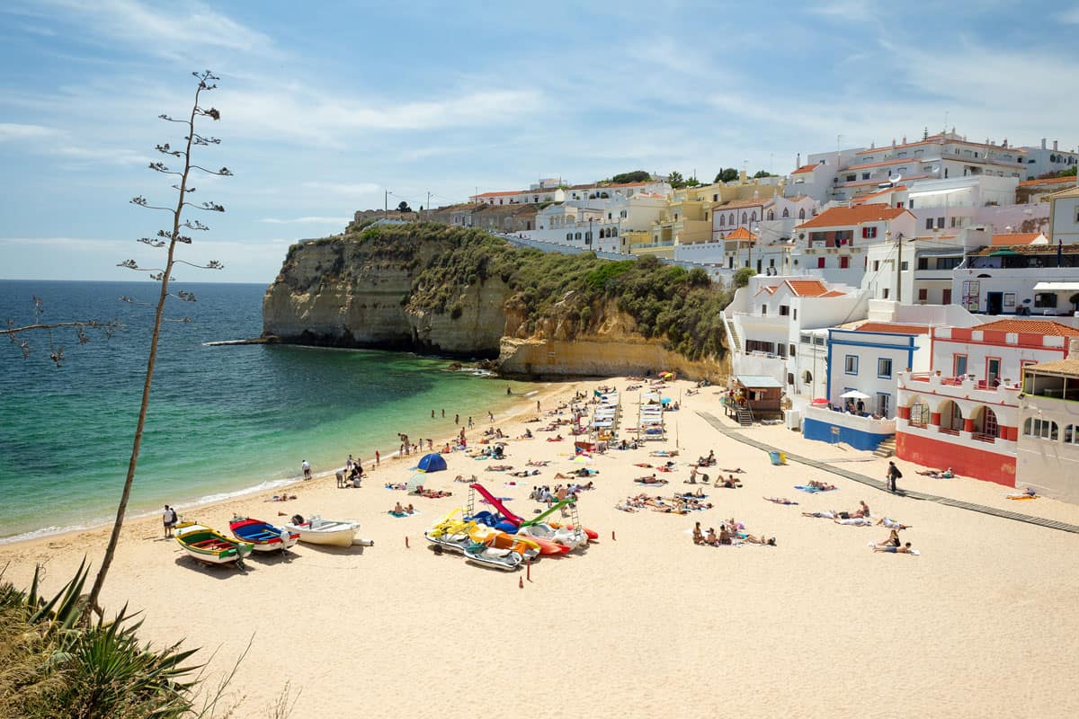 Traditional white Portuguese houses with red roofs line the cliffs all the the way down to the sand on the Beach of Carvoeiro where people sunbathe on the white sand and swim in the clear water on a sunny day. 