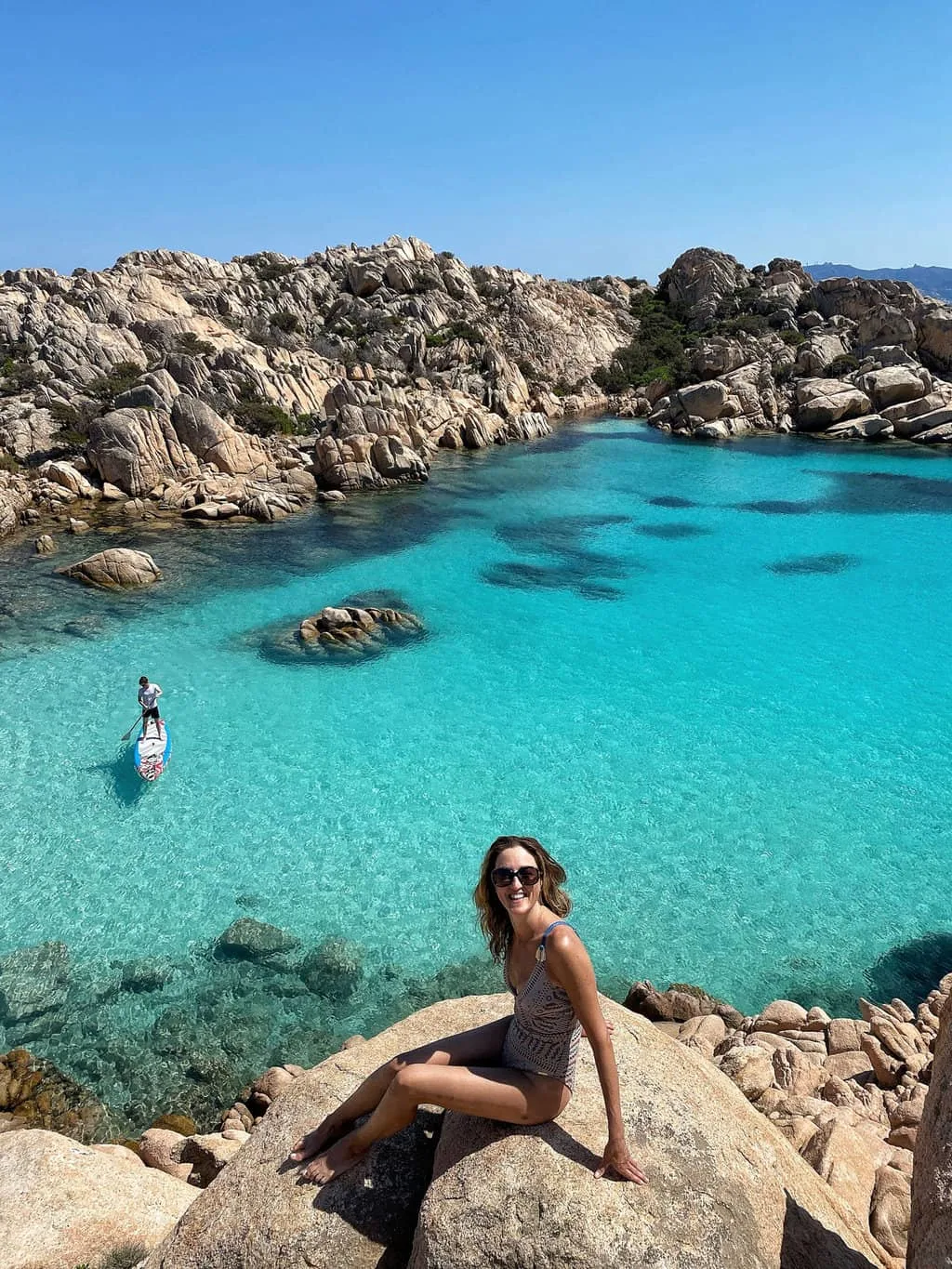 Young girl sitting on a rock in front  of the beach. A man is doing stand up paddle boarding on crytal clear water in the bay behind her in Cala Coticcio on Capera Italy. 