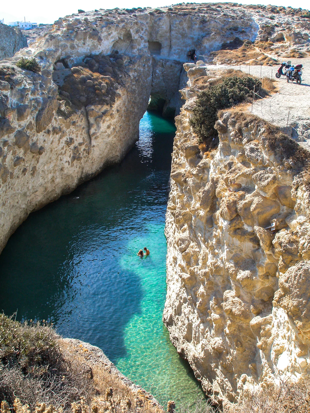 A couple swim in clear waters of Milos island surrounded by cliffs in an enclosed bay.