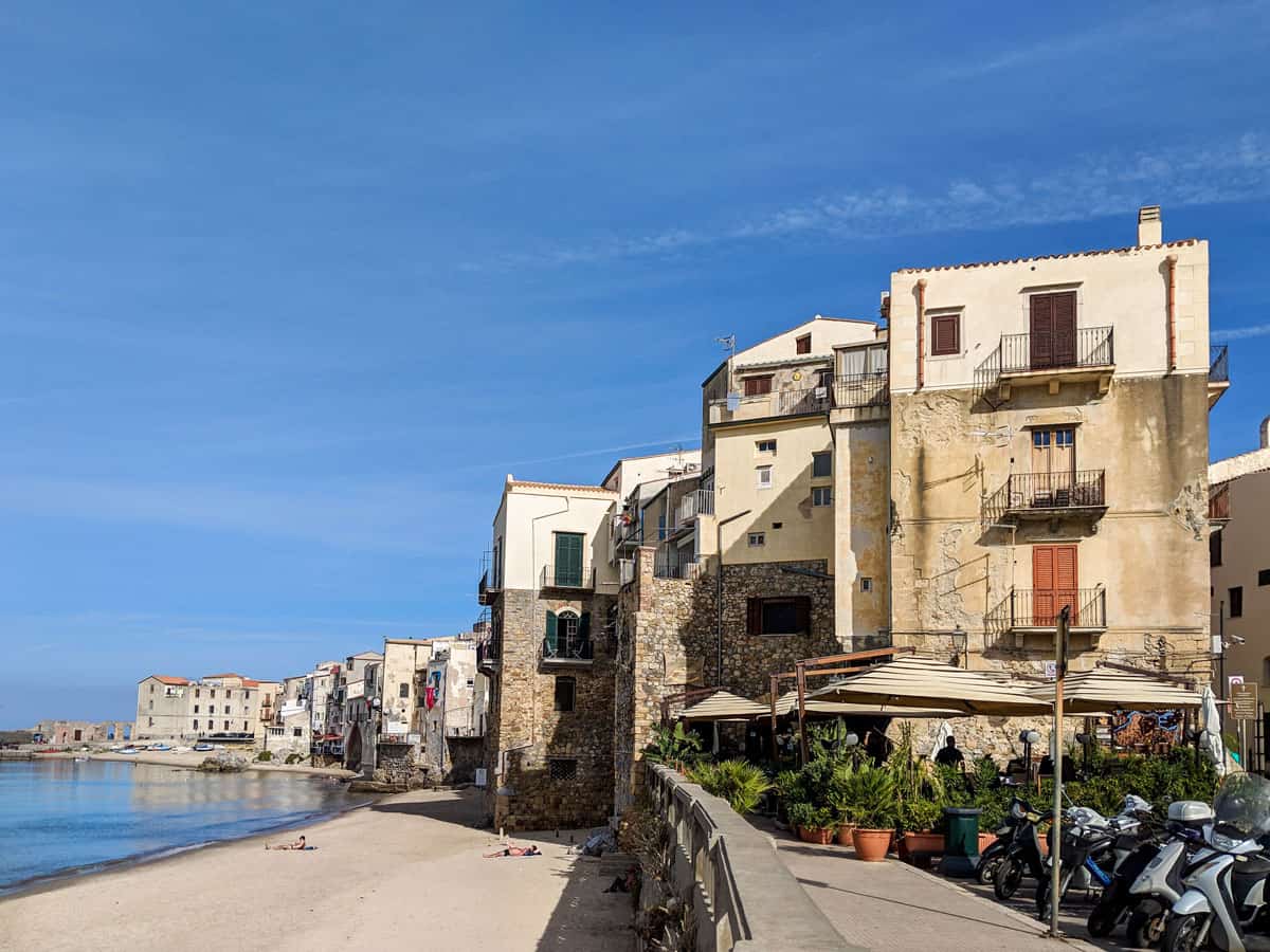 Two people lie on a white beach in front of typical stone buildings overlooking a calm sea in Cefalu Sicily. 