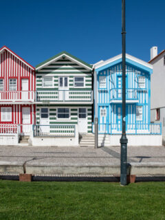 A tidy row of beach houses painted with candy stripes in red and white, blue and white and yellow and white stand along a pedestrian promenade edged with neat green grass.