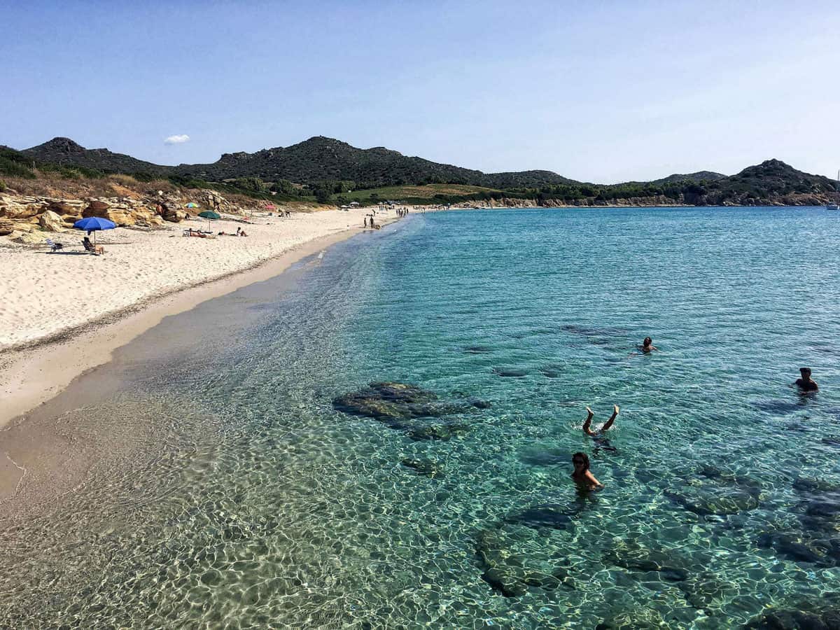 People swimming in clear water on a white sand beach in Sardinia Italy. 