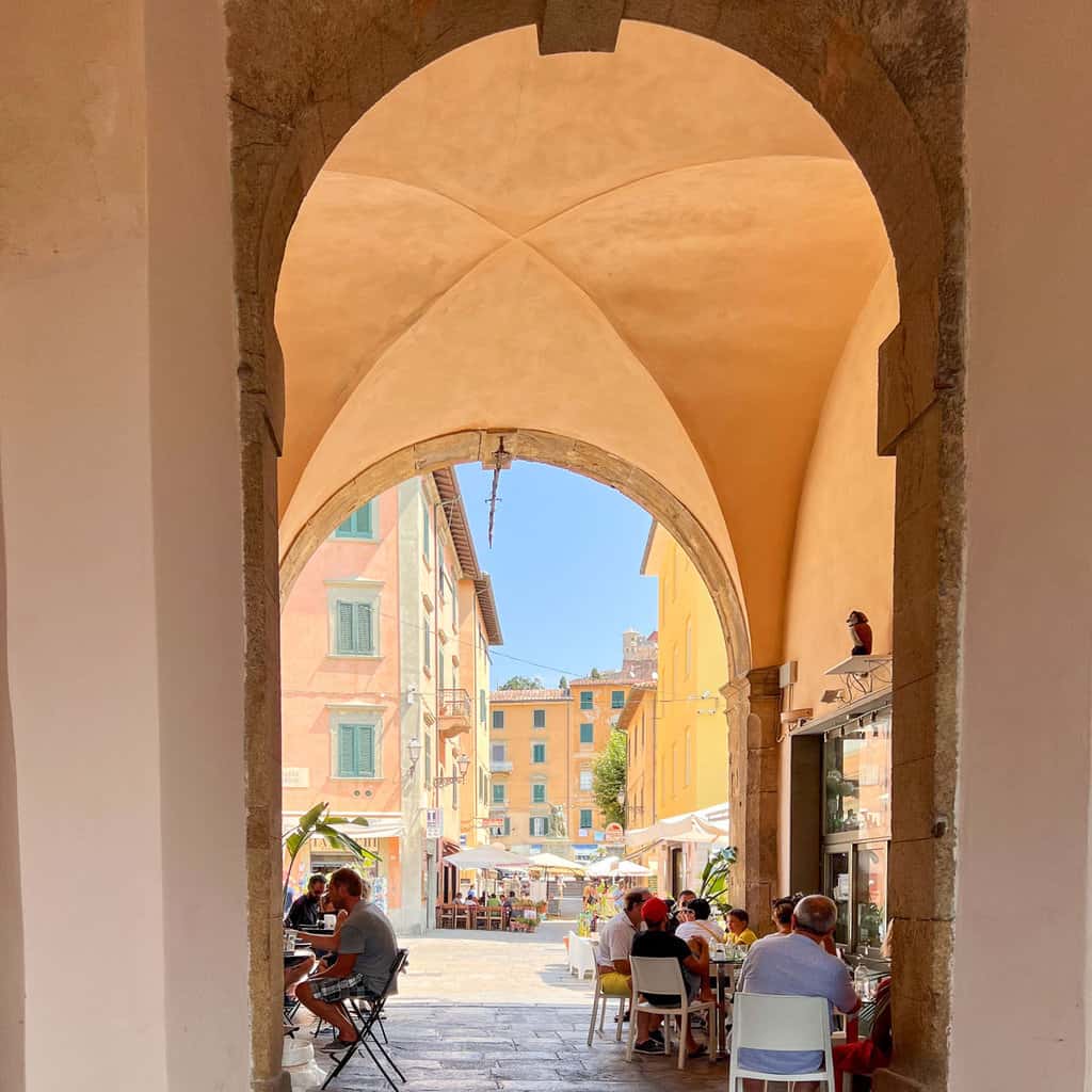 A stone arched walkway lined with cafe tables leading to a plaza in the old town of Elba Island.