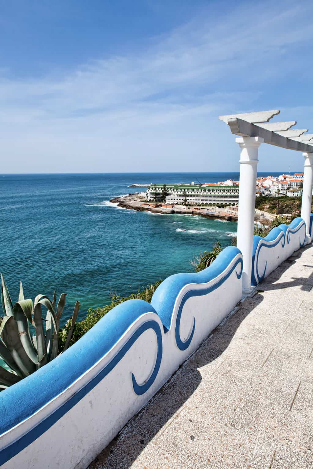 Blue and white decorative railings overlooking clear blue, calm ocean and the coastal surf town of Ericeira on a sunny day. 