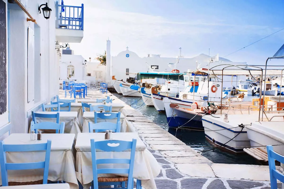 Tables with white tablecloths line the waterfront with traditional fishing boats moored in front. 
