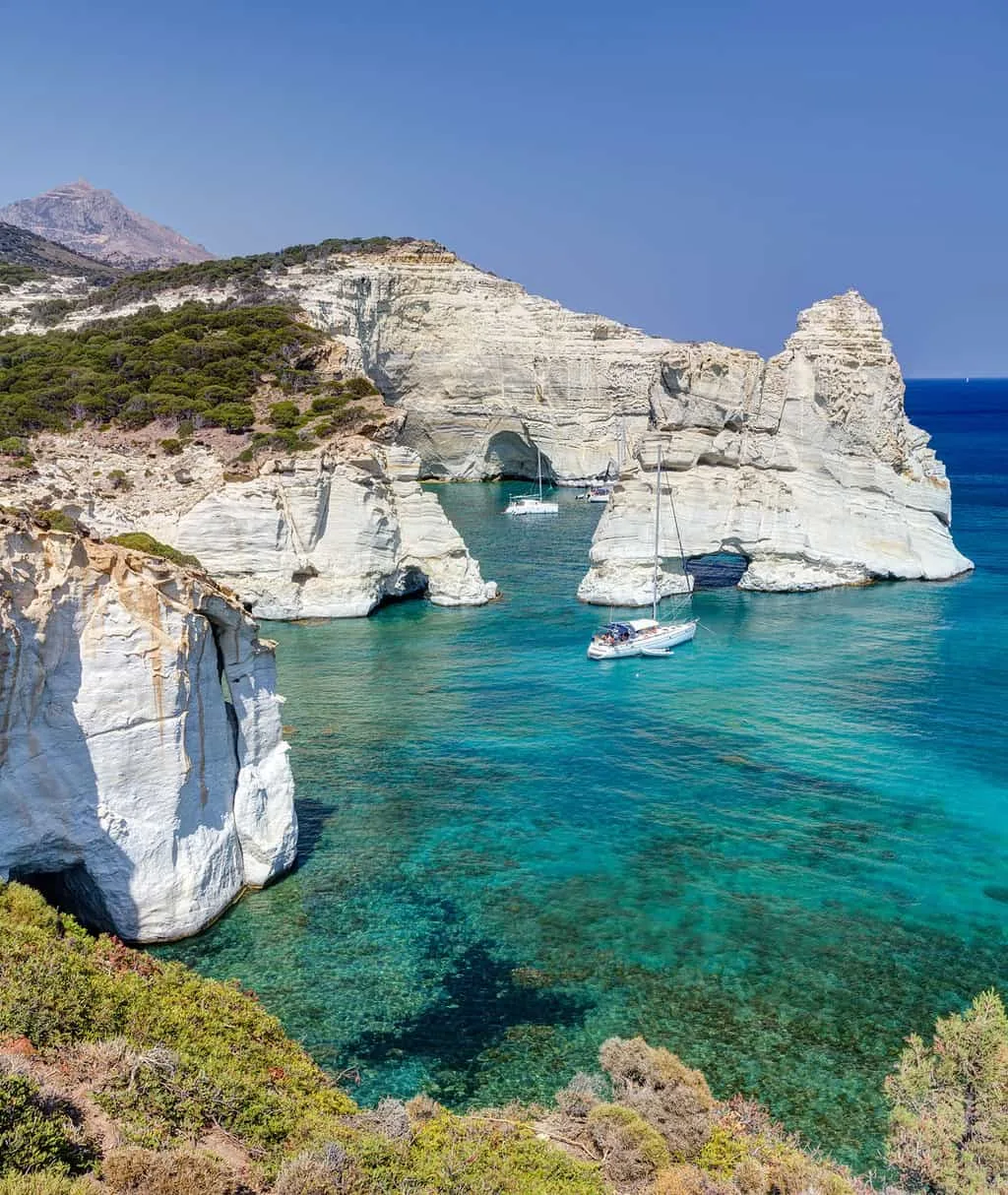 Boats anchored in crystal clear water surrounded by white cliffs on Milos Island Greece.