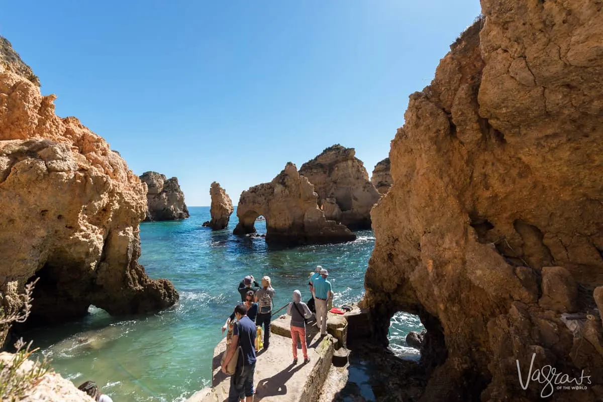 Tourists waiting on a small dock on a sunny day next to the clear, calm sea among the incredible coastal rock formations in Lagos Portugal. 