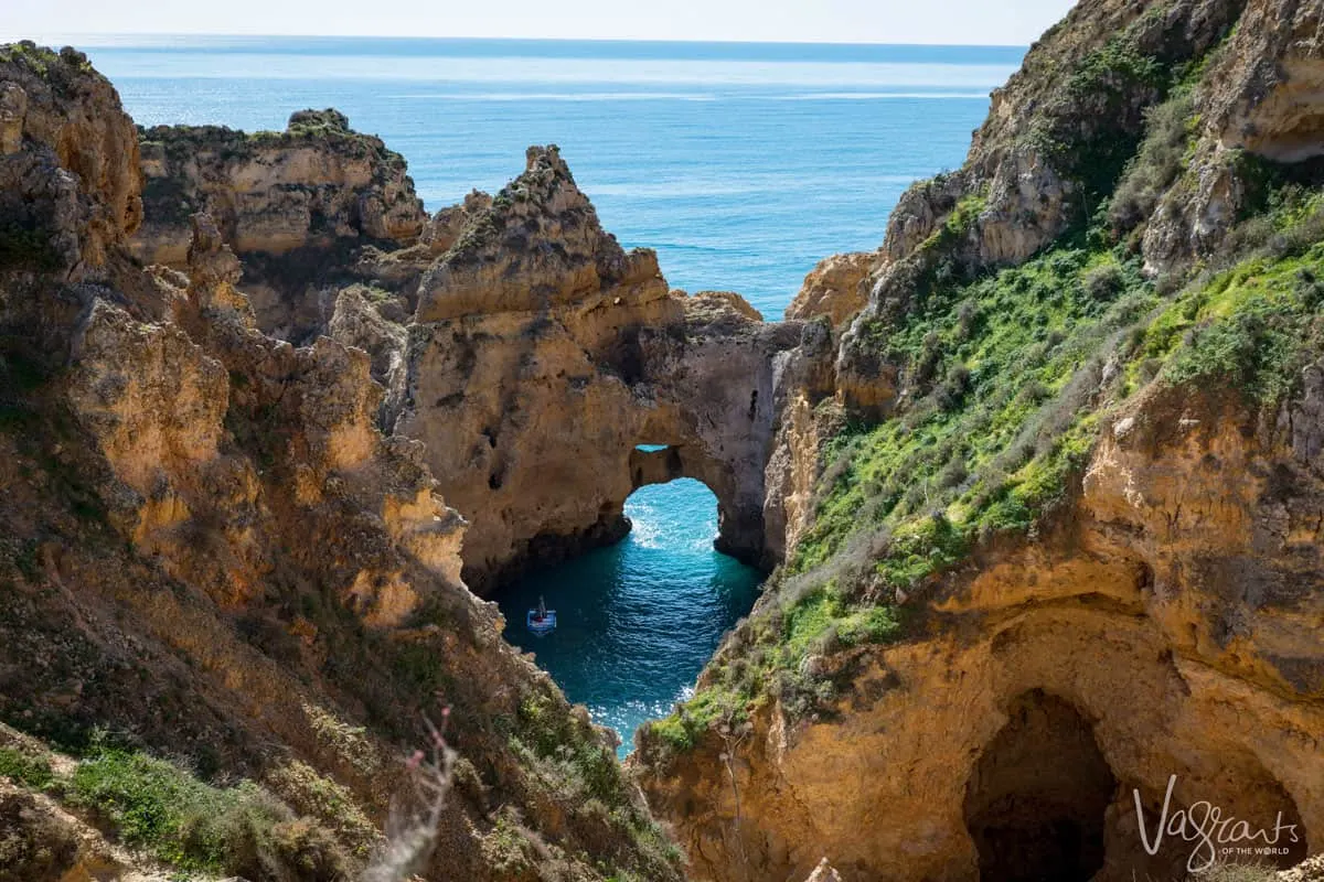 Yellow cliffs covered in bright green plants form a circular open cave around a tourist boat in clear calm water on the coast of Lagos Portugal. 