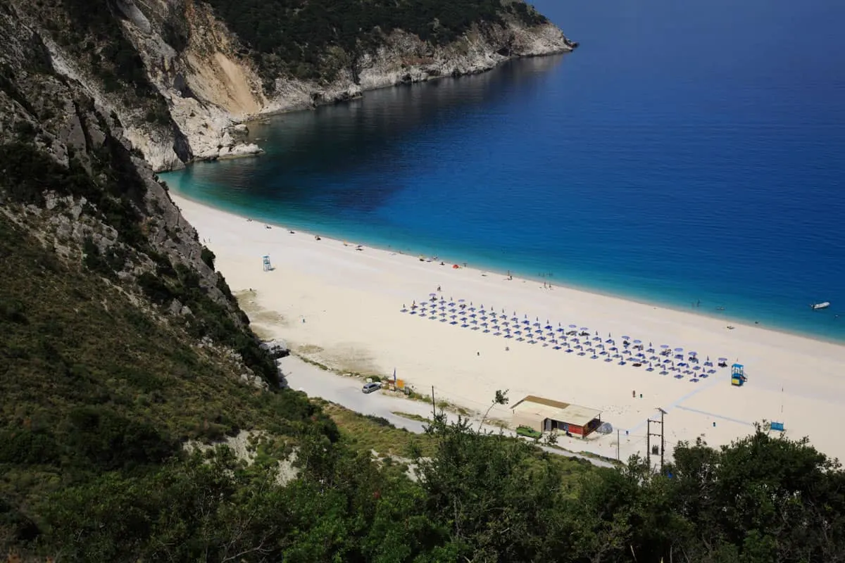 Looking over cliffs onto a white sand beach and deep blue water. There are neat rows of beach chairs on the beach.