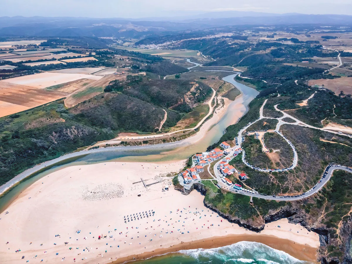 Aerial view of Praia de Odeceixe along Ribeira de Seixe river with people on the beach in summertime, and farm land in the distance. 
