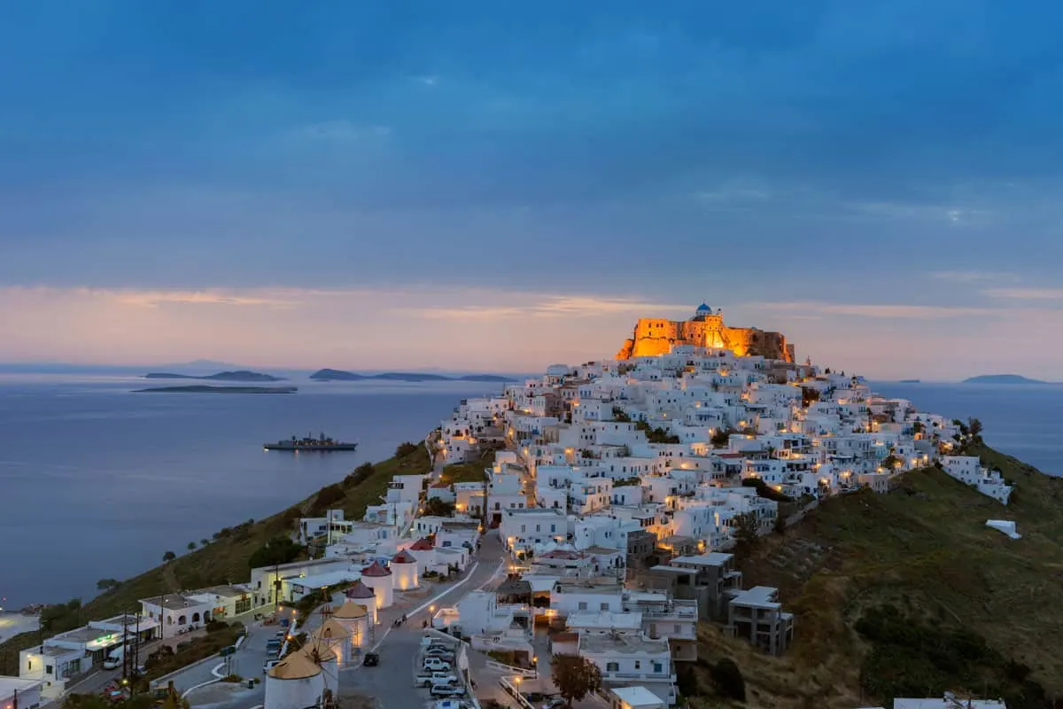 A castle sits on top of a hill still lit up in the low light of dawn. A white Greek village spills down the hill from the castle. 