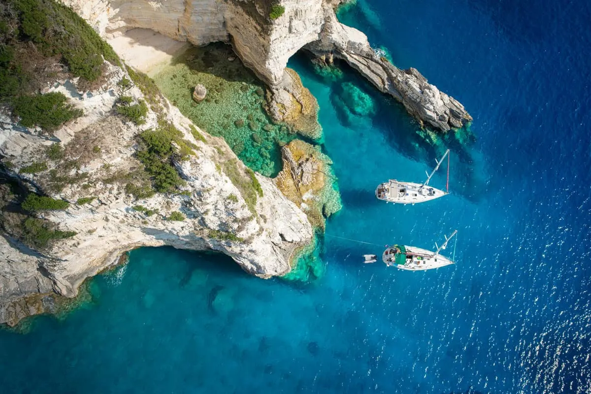 Aerial view of two yachts moored next to rugged white cliffs in clear turquoise water in Greece.