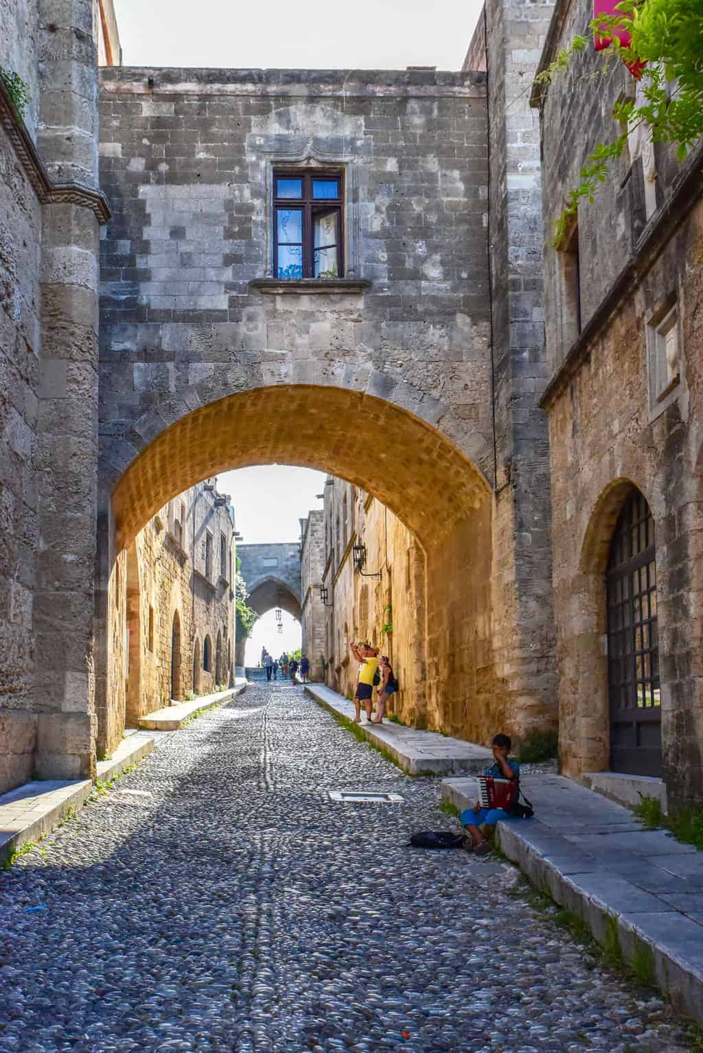 The cobbled streets of the Old Town in Rhodes with an archway over the street. Tourists are taking photos on the side of the street and a young boy with an accordion sits on the side of the road.  