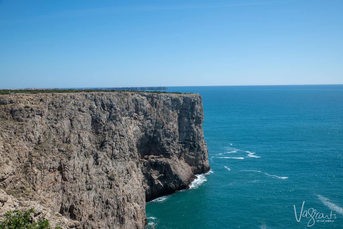 People stand on top of sheer cliffs that fall into a calm blue sea on the coast of Sagres in Portugal. The people are very small showing the immense size of the cliffs. 