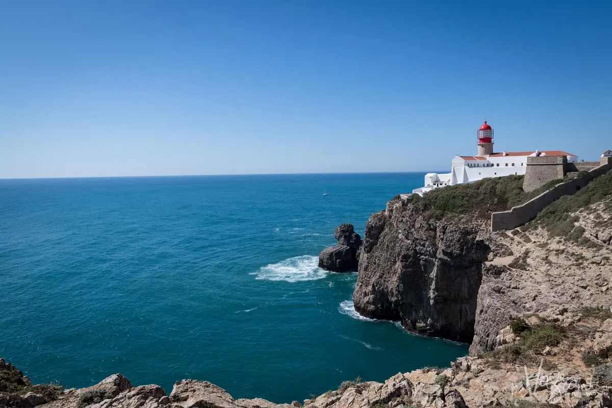 The red and white Cabo São Vincente lighthouse sits on the top of sheer cliffs that fall into a calm blue sea in Sagres Portugal. It is a clear, sunny day and a lone yacht sails in the distance. 