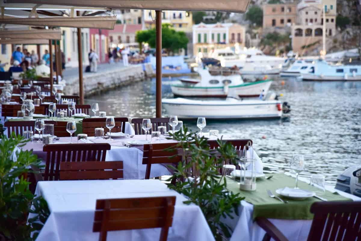Restaurant tables with white table cloths and wine glasses line the waterfront in Symi island Greece. Fishing boats are moored in the background. 