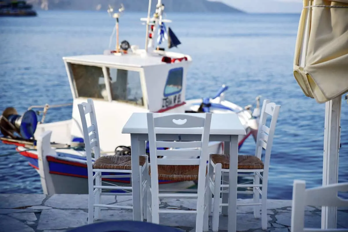 A white cafe table with chairs on the waterfront in Symi Island. A typical Greek fishing boat is moored on the blue water in front of the table. 